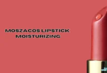 The image shows a red lipstick with a smooth, shiny surface, standing upright on the right side. The background is a matching red color. The text "MOSZACOS LIPSTICK MOISTURIZING" is centered on the left side of the image in bold, black, and white-outlined capital letters. At the bottom of the lipstick, there is a small logo with the words "THE RATINGS GURU."