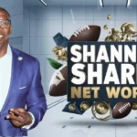 This image features Shannon Sharpe, a former NFL player and well-known sports analyst, wearing a purple suit and glasses, smiling confidently. He is making a double peace sign gesture with both hands. Next to him is a bold graphic with the text "Shannon Sharpe Net Worth" in large white and gold letters. Surrounding the text are various symbols, including footballs, gold coins, a microphone, and an upward arrow on a bar chart, all symbolizing his financial success, media influence, and career in sports. The background is sleek and modern, with a professional aesthetic.