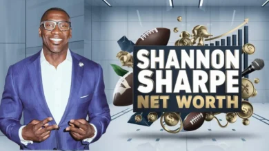 This image features Shannon Sharpe, a former NFL player and well-known sports analyst, wearing a purple suit and glasses, smiling confidently. He is making a double peace sign gesture with both hands. Next to him is a bold graphic with the text "Shannon Sharpe Net Worth" in large white and gold letters. Surrounding the text are various symbols, including footballs, gold coins, a microphone, and an upward arrow on a bar chart, all symbolizing his financial success, media influence, and career in sports. The background is sleek and modern, with a professional aesthetic.