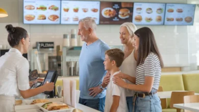 A family of four stands in line at a modern fast-food counter, ready to place their order. The father is at the front, speaking with the cashier, while the mother stands behind with a hand on the shoulder of their young son. Their teenage daughter is next to them, waiting her turn. The scene is bright and welcoming, with digital menu screens above displaying images of various food options. The setting is clean and contemporary, with a casual, friendly atmosphere.