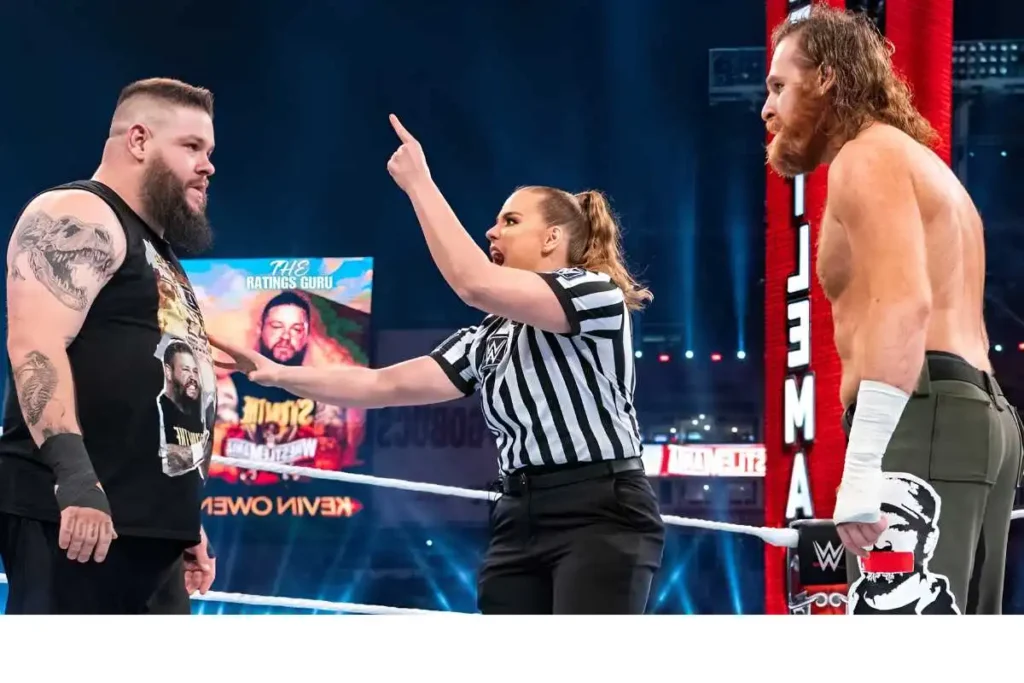 In-ring scene from WWE SmackDown Episode 1491 showing two wrestlers in an intense faceoff, with a female referee stepping in to separate them. The wrestler on the left, sporting a beard and black sleeveless shirt with visible tattoos, stares down his opponent on the right, who is dressed in green pants and has white tape on his hand. The background features a large screen with 'The Ratings Guru' watermark and other event graphics, adding to the high-energy atmosphere of the moment.